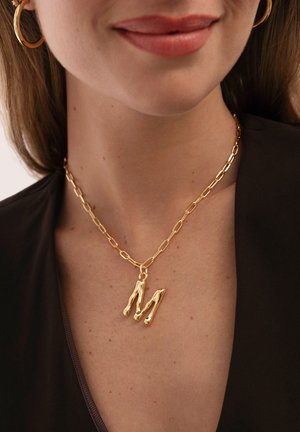 Mujer sonriente con pendientes de aro dorados y un collar de cadena dorado con un colgante grande en forma de letra M sobre una blusa negra.