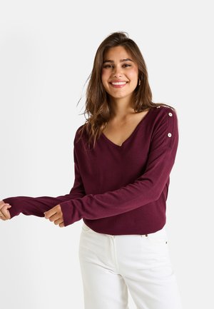 Pull rouge bordeaux à manches longues avec un col en V, orné de boutons décoratifs sur l'épaule et de poignets côtel�és. Porté avec un pantalon blanc.