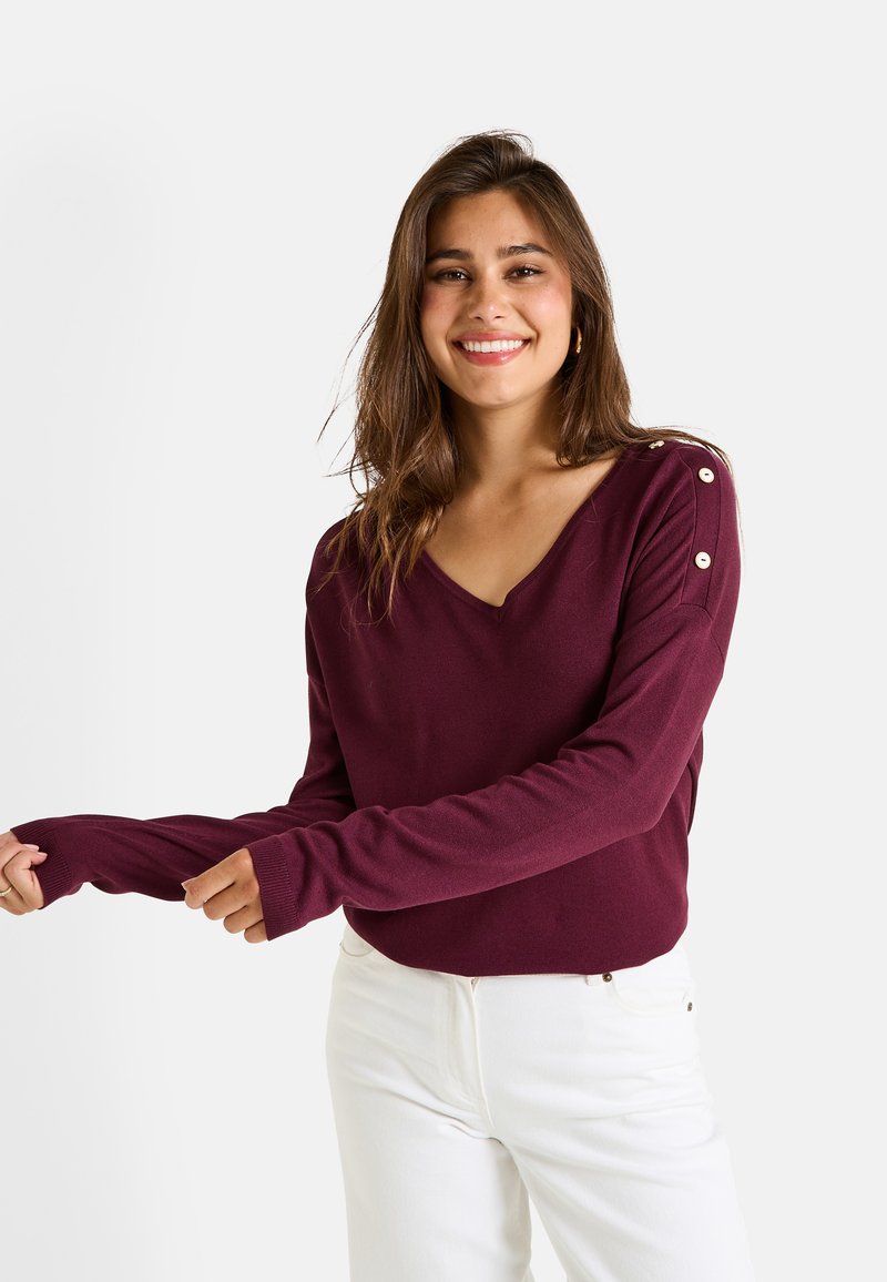 Pull rouge bordeaux à manches longues avec un col en V, orné de boutons décoratifs sur l'épaule et de poignets côtelés. Porté avec un pantalon blanc.
