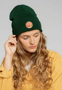 Zielona dziana czapka z podwiniętym mankietem i okrągłą tanową skórzaną łatką z motywami górskimi i zwierzęcymi.