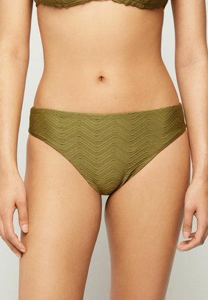 Mujer con braguita de bikini verde oliva texturizada con patrón ondulado, mostrando el torso y la parte superior de los muslos sobre un fondo liso.