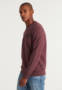 Sudadera burdeos con cuello redondo, puños y dobladillo acanalados; presenta un pequeño parche de logo negro en el pecho. Combinada con unos vaqueros azul claro.