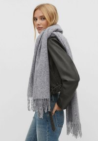 Écharpe grise en tricot avec des franges, posée sur une veste en cuir noire, portée avec un jean en denim bleu. Texture douce avec un motif subtil.