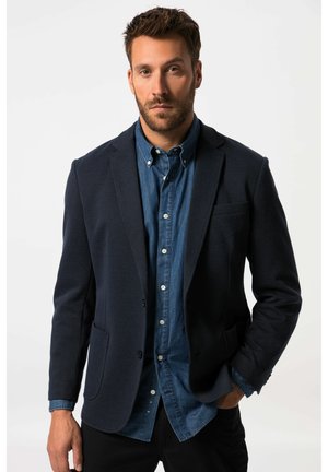 Mand med kort brunt hår og skæg iført en mørk blazer over en blå skjorte med knapper, står med den ene hånd i lommen mod en hvid baggrund.