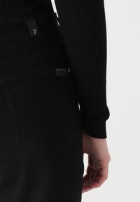 Zwarte denim broek van een textuurstof, met achterzakdetail en een zwart geribd langemouwen top dat gedeeltelijk zichtbaar is op de bovenrug.