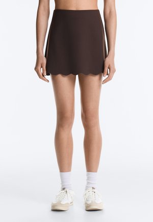 Žena v tmavohnedej minisukni so zvlneným lemom, bielych ponožkách a bielych teniskách, stojaca pred jednofarebným pozadím.