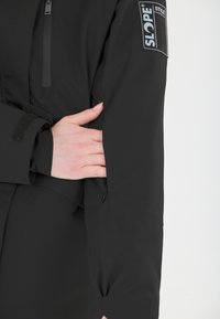 Musta ulkovaatteet, joissa on vetoketjullinen tasku, säädettävä hihansuu ja SLOPE STYLE -logopatchi hihassa. Sujuva pinta.