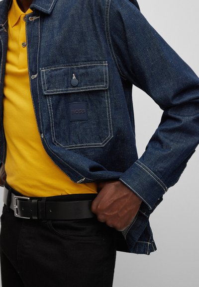 Veste en denim de couleur bleu foncé, avec des coutures visibles, une poche à rabat et un logo. Portée par-dessus un polo jaune et un jean noir avec une ceinture.