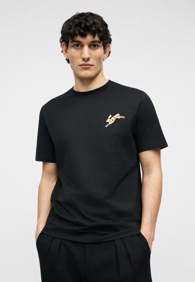 Νέος άνδρας με κοντά σγουρά μαλλιά, φοράει μαύρο T-shirt με μικρό καφέ λογότυπο λαγού στο στήθος, με τα χέρια στις τσέπες, ουδέτερο φόντο.