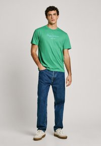 Žalias medvilninis marškinėliai su apvaliu iškirpimu ir baltu "Pepe Jeans London" logotipu, derinami su mėlynomis džinsinėmis kelnėmis ir baltomis bateliais.