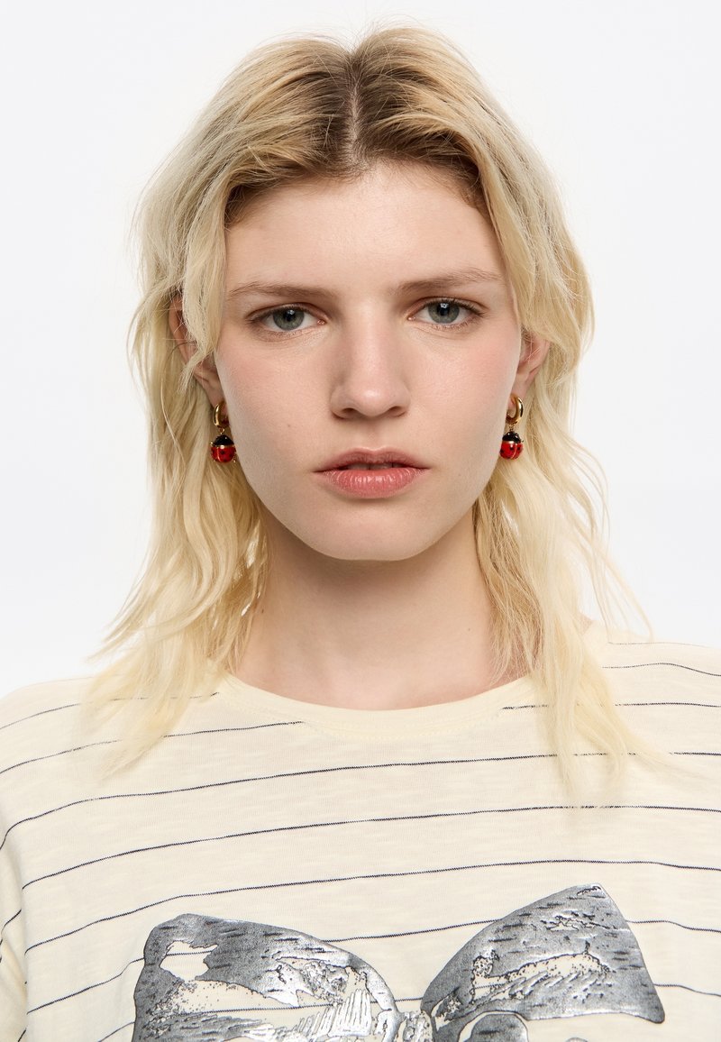 T-shirt a righe giallo chiaro con una grafica a forma di fiocco metallico argento. La modella indossa orecchini a forma di coccinella rossa, ha i capelli biondi ondulati e trucco naturale.