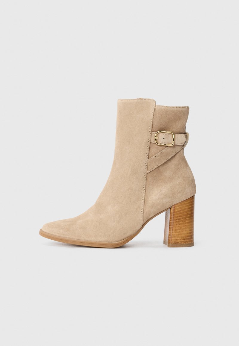 Stivaletto in suede beige con punta affusolata, tacco a blocco di altezza media e un elemento decorativo in metallo dorato sulla parte laterale.