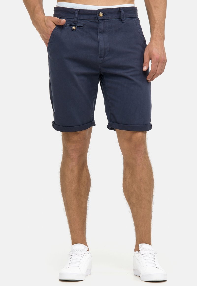 Homme portant un short bleu marine retroussé et des baskets blanches, debout avec la main gauche dans la poche, sur un fond uni.