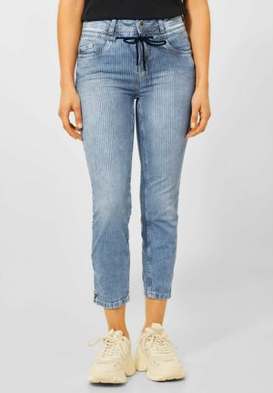 Frau trägt hellblaue, vertikal gestreifte, kurz geschnittene Jeans mit schwarzem Kordelzug und klobigen, cremefarbenen Sneakern.