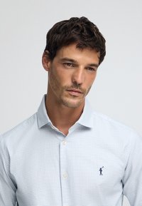 Camicia a quadretti blu chiaro con colletto button-down. Presenta un piccolo logo scuro sul lato sinistro del petto. Realizzata in un tessuto liscio.