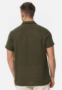Camicia a maniche corte di colore verde scuro, in materiale testurizzato. Presenta un colletto classico e una linea dritta con un orlo posteriore leggermente più lungo.