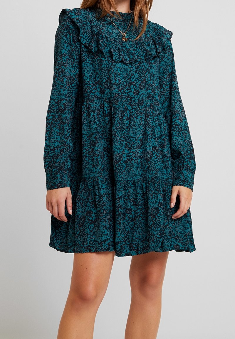 Femme portant une mini-robe à manches longues bleu sarcelle et noire, avec col à volants et jupe à volants, debout devant un fond uni.