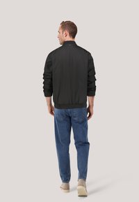 Schwarze Bomberjacke mit gerippten Bündchen und Kragen, mit glatter Textur. Trägt sie über einer blauen Jeans und beigen Schuhen, von hinten gesehen.