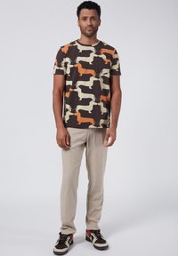 Brun t-shirt med ett mönster av taxar i kräm, orange och brunt. Matcher med beige byxor och mångfärgade sneakers.