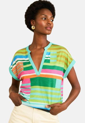 Kvinde med kort krøllet hår iført guldringe og en flerfarvet stribet skjorte med V-hals og opkrogede ærmer, hænderne i lommerne.