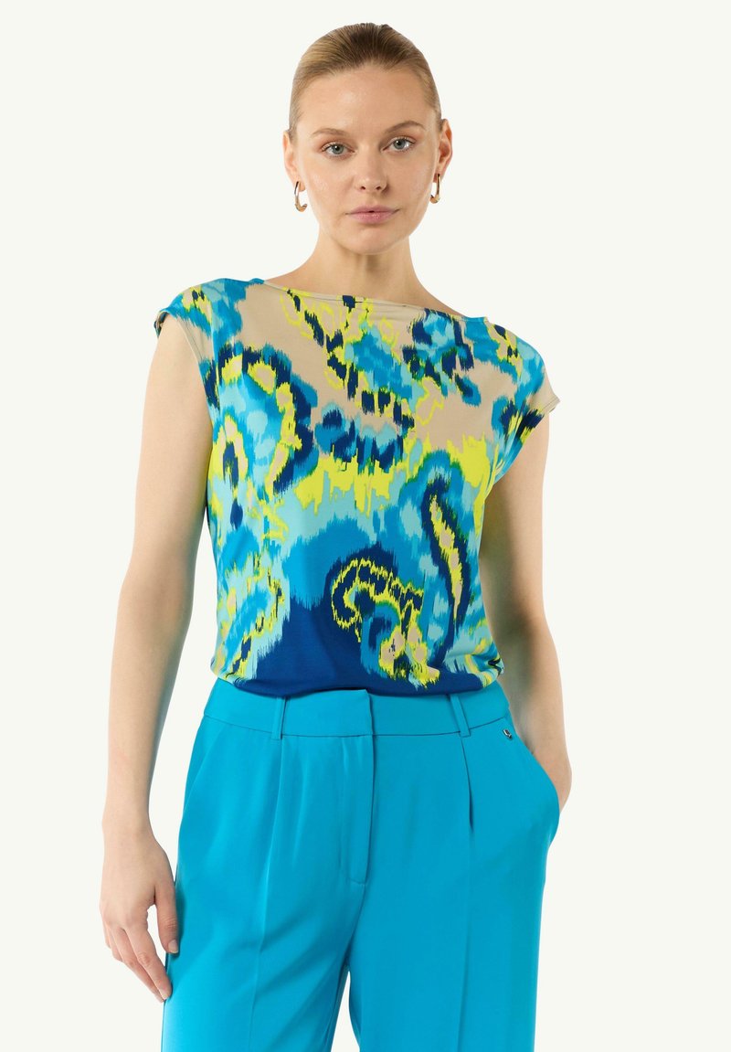 Korte-mouwen top met een multikleurig abstract patroon in blauw, groen en geel; gecombineerd met turkooise broek. Gladde stof structuur.