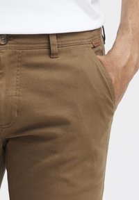 Pantalones caqui hechos de una tela suave, con un corte clásico, lazos para cinturón y una etiqueta de cuero en la cintura. Bolsillo delantero visible.