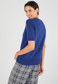 Pull bleu à manches courtes avec une texture douce, avec des poignets et un ourlet côtelés, associé à un pantalon à carreaux gris et bleu.