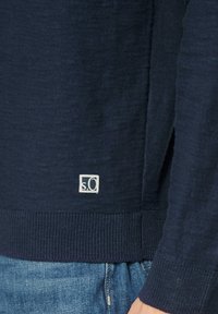 Granatowy sweter z teksturowanym splotem, z rzebrowanym dołem i mankietami. Mała naszywka z logo w kwadratowym kształcie blisko dolnej krawędzi.
