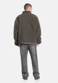 Kurtka z polaru w ciemnym oliwkowym zielonym kolorze z wysokim kołnierzem i teksturowaną powierzchnią, zestawiona z szarymi spodniami dżinsowymi i czarnym obuwiem.