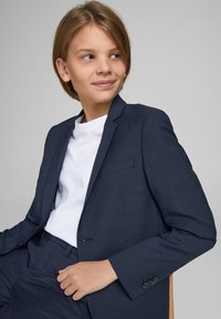 Costume bleu marine avec une texture subtile et un design ajusté, comprenant une veste à un bouton et un pantalon assorti, porté sur un t-shirt blanc.