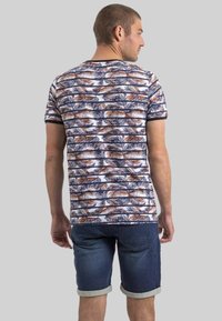 T-shirt à manches courtes en blanc avec un motif de feuilles de palmier bleu marine et rouille ; associé à un short en denim foncé, avec des roulettes aux manches et une coupe décontractée.