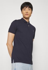 Mann steht vor weißem Hintergrund, trägt ein marineblaues Polohemd und hellblaue Jeans und schaut leicht nach oben und nach vorne.