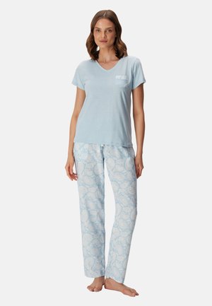 Vrouw staat op blote voeten, draagt een lichtblauw topje met korte mouwen en een blauw-witte pyjamabroek met patroon tegen een effen achtergrond.