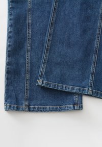 Tessuto denim blu scuro con taglio a gamba dritta, dettagli di cuciture visibili e orlo leggermente sfilacciato nella parte inferiore.