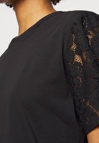 Camiseta negra con mangas cortas, hecha de intrincada encaje negro con patrones florales, de textura suave y diseño clásico de cuello redondo.