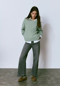 Joven mujer vistiendo un suéter de punto trenzado verde claro sobre una camisa blanca, jeans grises y zapatos mocasines marrones, de pie contra una pared lisa.