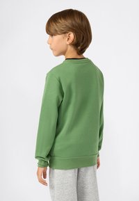 Enfant aux cheveux châtain clair portant un t-shirt à manches longues vert et un pantalon gris, tourné vers la gauche sur un fond blanc uni.