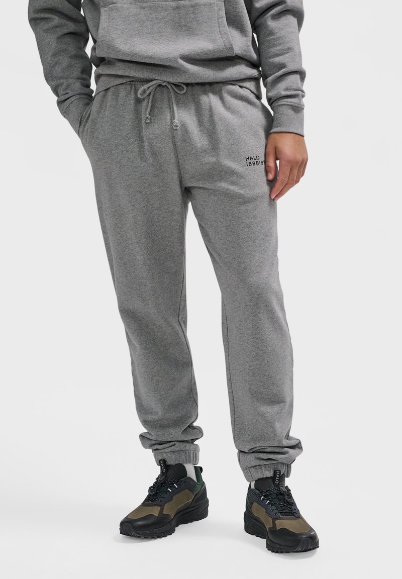 Mand iført grå joggingbukser med teksten "HALO 196819" på låret, kombineret med sorte og grønne snøresneakers.