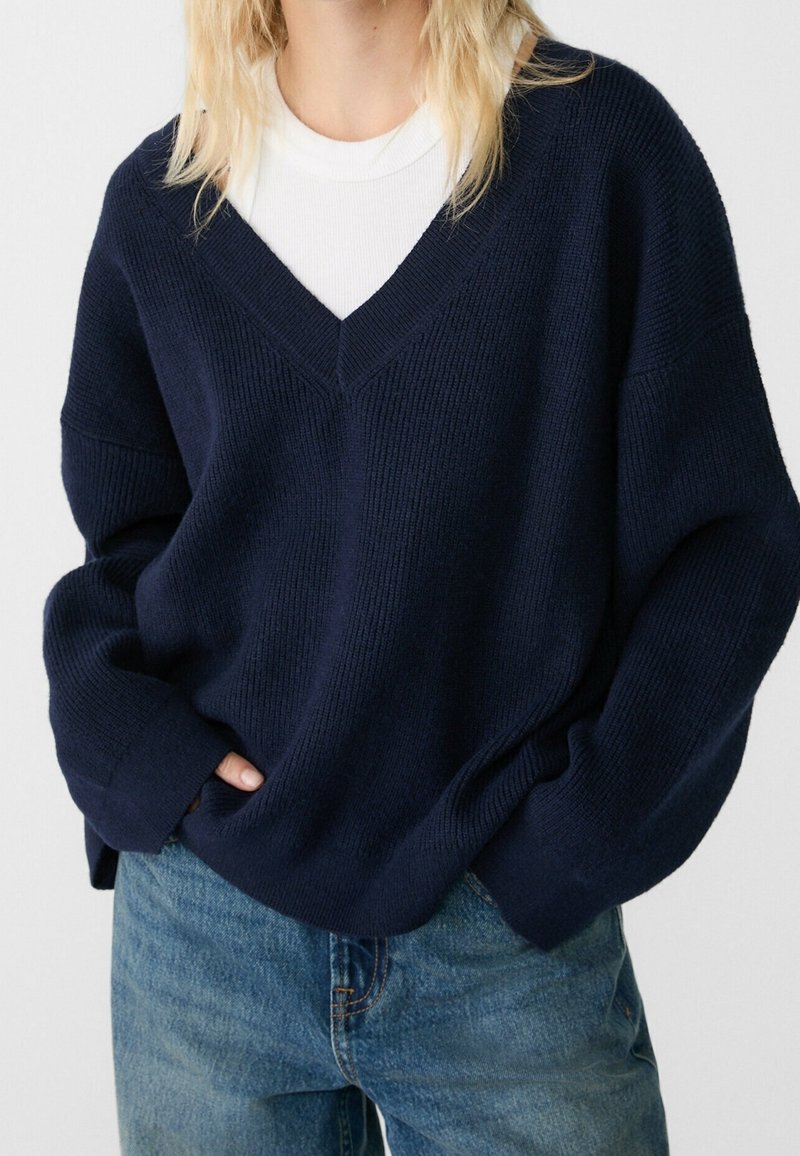 Personne portant un pull bleu marine à col en V sur une chemise blanche, associé à un jean en denim bleu, les mains dans les poches.