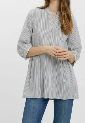 Kobieta w luźnej bluzce w pionowe paski w kolorze szaro-białym z długimi rękawami i dekoltem w kształcie litery V, w połączeniu z niebieskimi dżinsami na jednolitym tle.