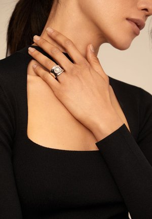 Anillo de plata con un detalle circular en el centro, llevado en una mano contra una blusa negra de manga larga con relieve. Vista en primer plano centrada en las manos y la joyería.