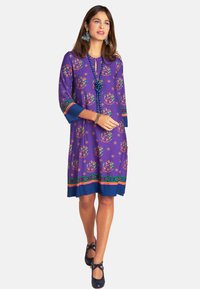 Lila Kleid mit Blumendrucken, Dreiviertelärmeln und einem bunten Saum. Kombiniert mit blauen Schuhen und dekorativen Ohrringen.