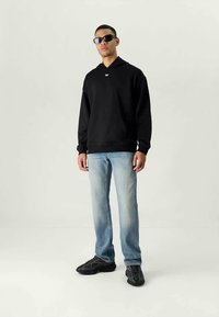 Homem em pé, vestindo um hoodie preto, jeans azul claro, sapatilhas pretas e óculos de sol escuros contra um fundo claro liso.