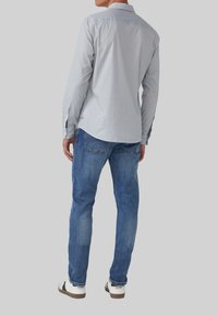 Mann trägt ein hellgraues Hemd mit Knöpfen, blaue Jeans und weiße Sneakers und steht mit dem Rücken zu einem schlichten grauen Hintergrund.