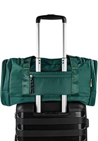 Bolsa de lona verde con asas dobles y bolsillo con cremallera, colocada sobre una maleta negra con ruedas y mango telescópico de aluminio.