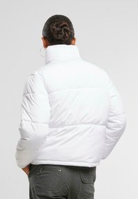 Bijela puffer jakna s visokim ovratnikom, quilted dizajn i elastične rukave, uparen s tamnosivim hlačama koje imaju detalj s patentnim zatvaračem.