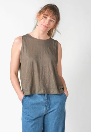 Žena svijetlosmeđe kose u okrutnom maslinastom topu bez rukava i plavim trapericama s visokim strukom s rukama u džepovima, na bijeloj pozadini.