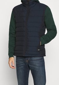 Granatowa pikowana kamizelka z zamkiem błyskawicznym i pikowanym wzorem, zestawiona z ciemnozielonym kapturem, noszona z niebieskimi dżinsami.