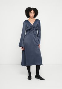 Rochie din satin bleumarin cu decolteu în V, mâneci lungi și detalii drapate în talie. Lungime până la genunchi, cu o textură netedă și fluidă. Pantofi negri cu talpă joasă.