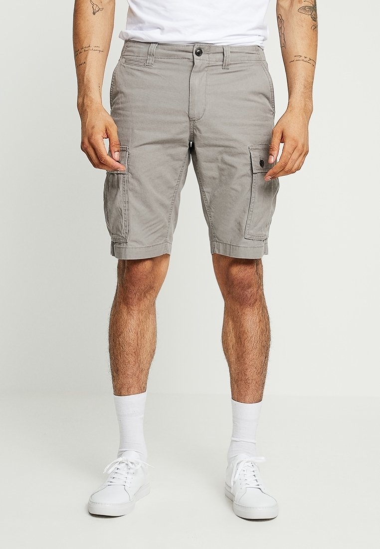 Homme portant un short cargo gris clair, des baskets blanches et des chaussettes blanches, debout devant un fond clair uni.